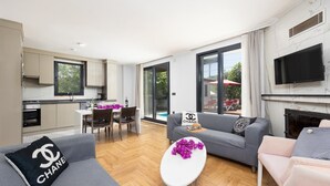 Ein modernes Wohnzimmer mit grauem Sofa, einem weißen Couchtisch und einem Essbereich mit Tisch und Stühlen. An der Wand ist ein Fernseher angebracht und es gibt einen Kamin.