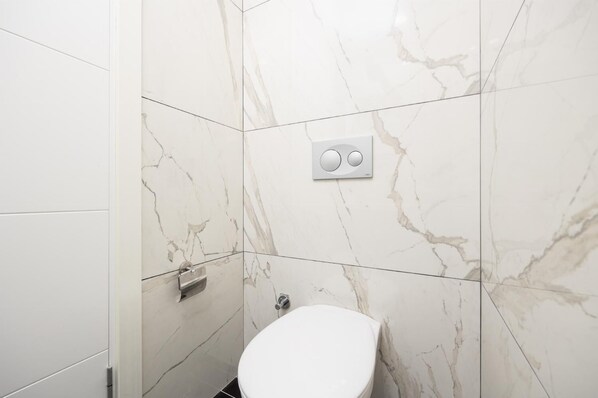 A bathroom with white marble tiles, a toilet, and a wall-mounted flush handle.