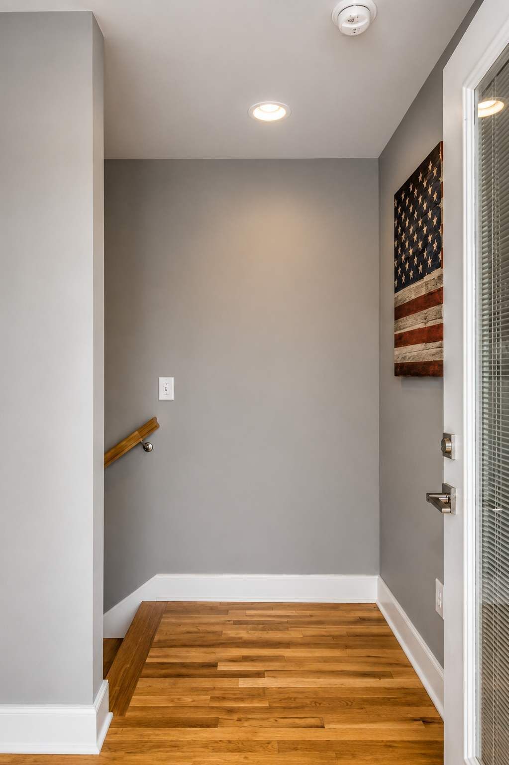 A hallway with a wooden floor, a gray wall, and a door with a window.