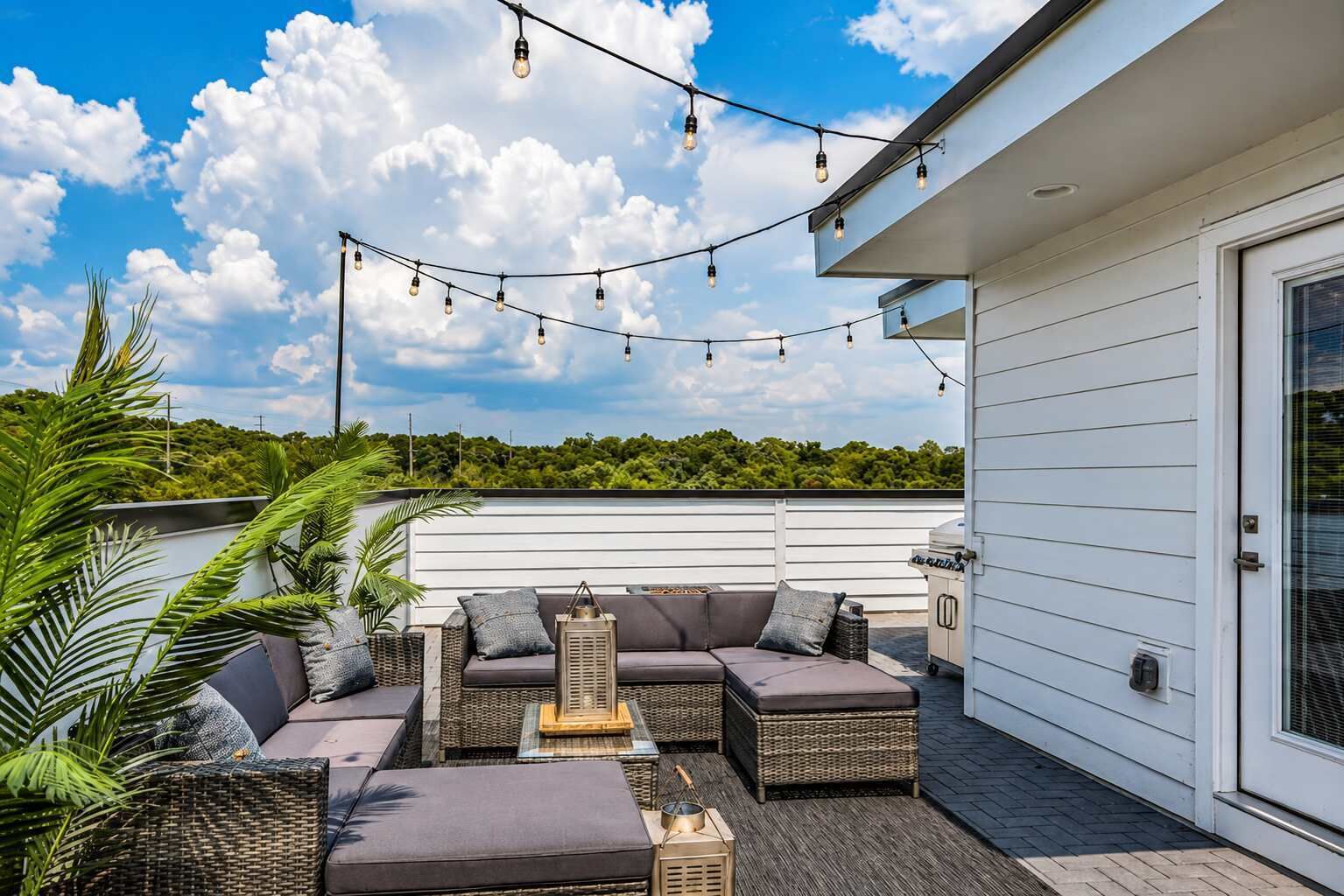 Eine Terrasse im Freien mit einer Sofagarnitur, Korbmöbeln und Lichterketten, vor einer Kulisse aus blauem Himmel und Grünpflanzen.