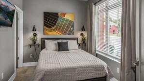 A bedroom with a bed, a painting, a window with blinds, and a view of a building outside.