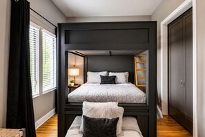 A bunk bed room with a desk and lamp, a window with blinds, and a wardrobe.