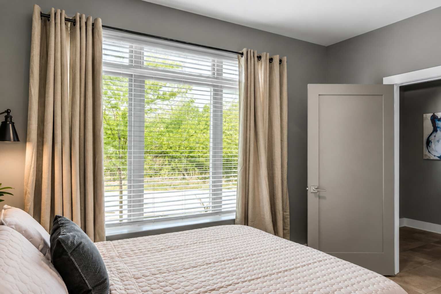 A bedroom with a bed, curtains, a window with blinds, a door, and a wall-mounted lamp.