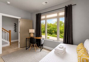 A bedroom with a bed, a desk, and a chair. There is a window with curtains, a door, and a staircase.
