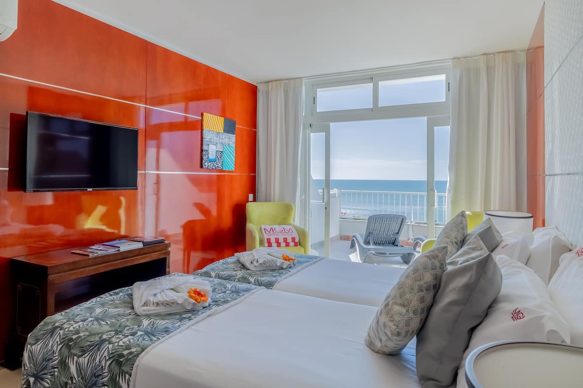 A hotel room with a large bed, a TV mounted on the wall, a balcony with a view of the sea, and a chair with a pillow labeled "MGM Grand Hotel".
