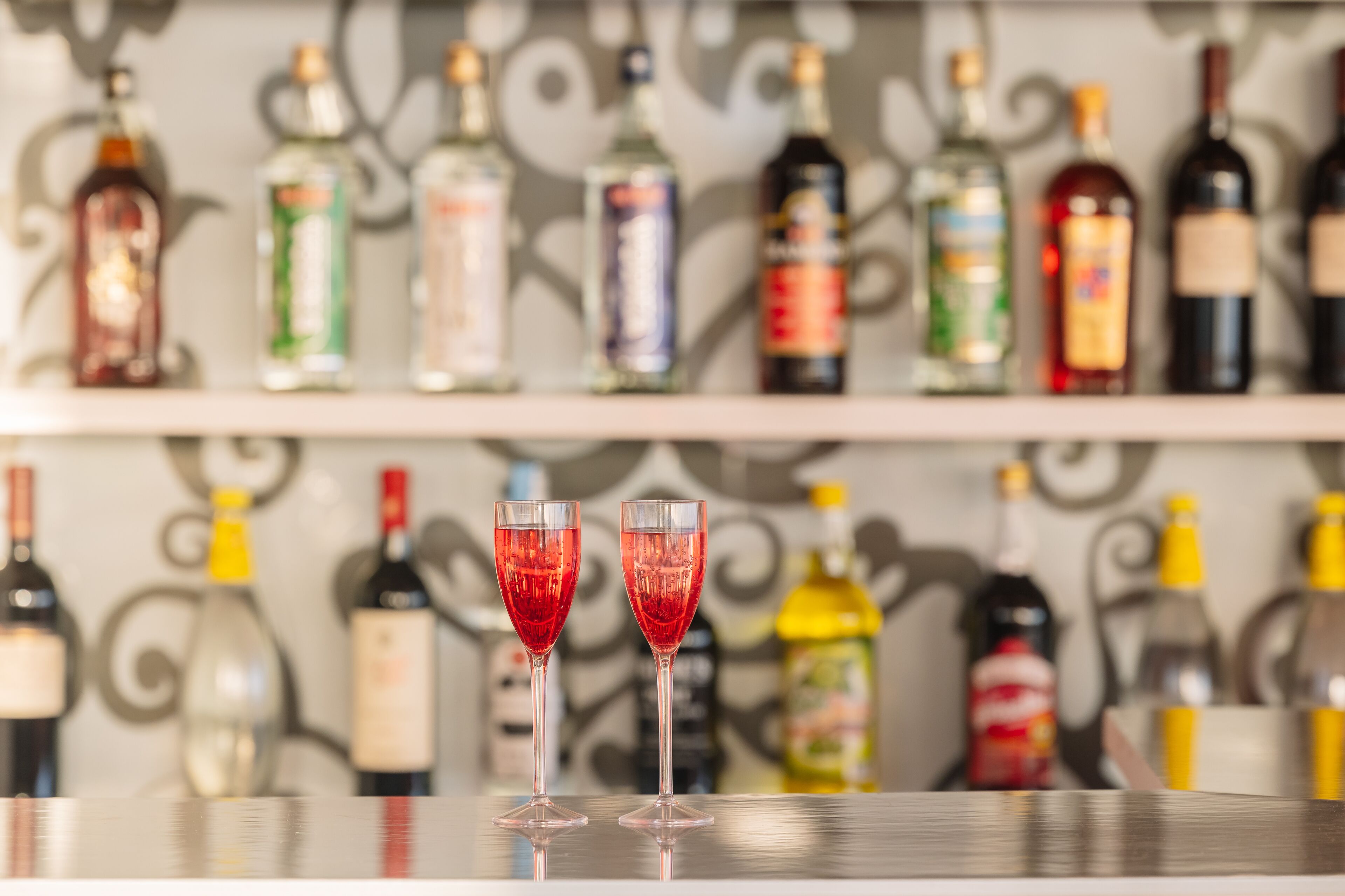 Dos vasos de cóctel rojo sobre una barra con una variedad de botellas de licor al fondo.