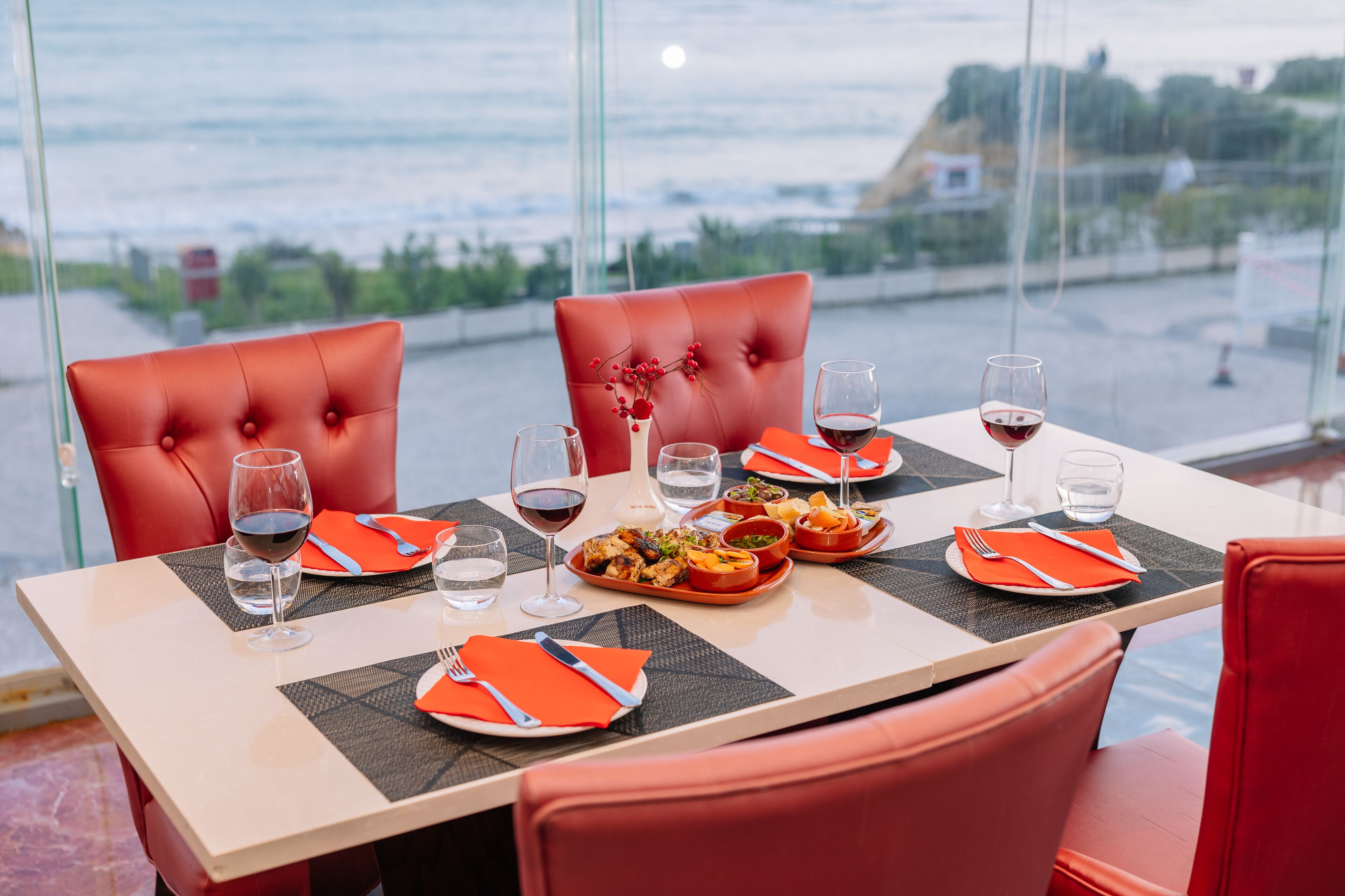 A dining table with red chairs, set with plates, cutlery, and glasses of red wine.