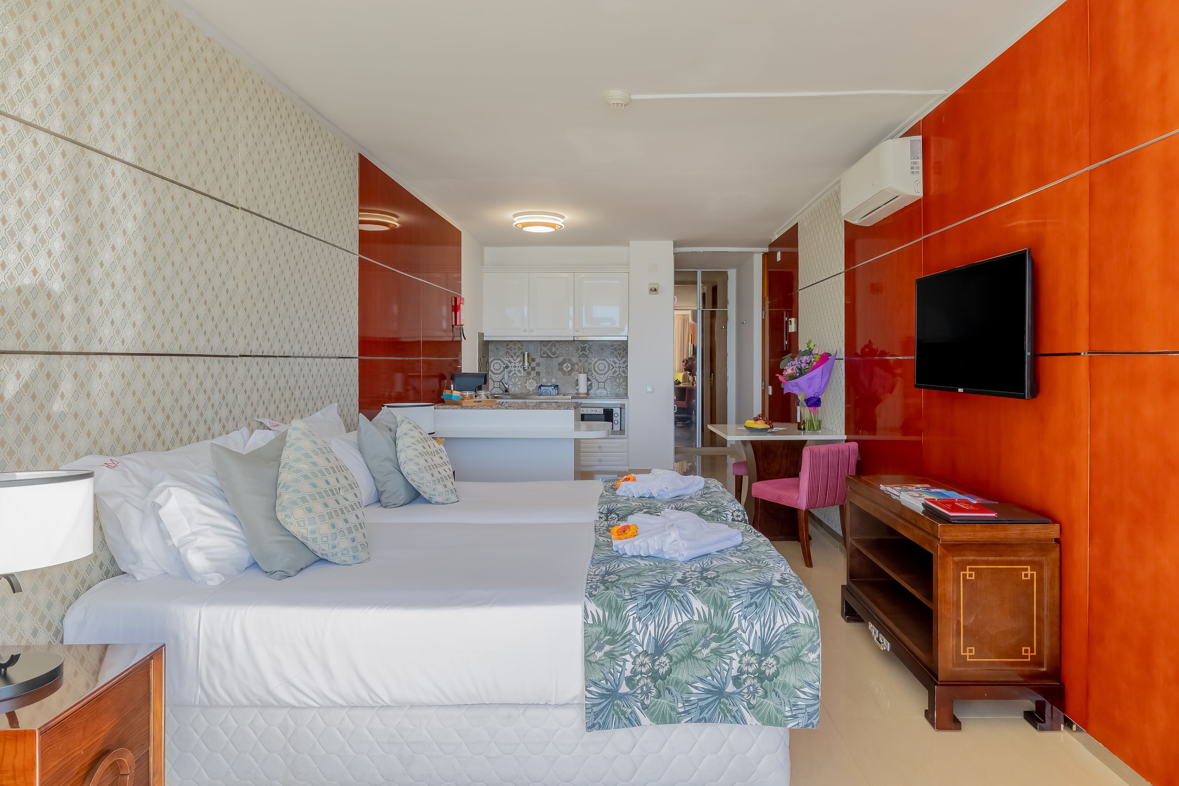 A hotel room with a bed, bedside table, TV, and a kitchenette area.