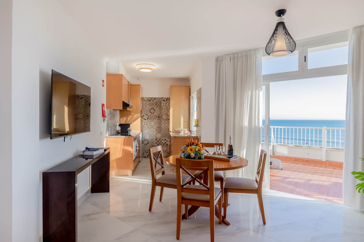 A modern living room with a dining area, a TV, and a balcony with a view of the sea.