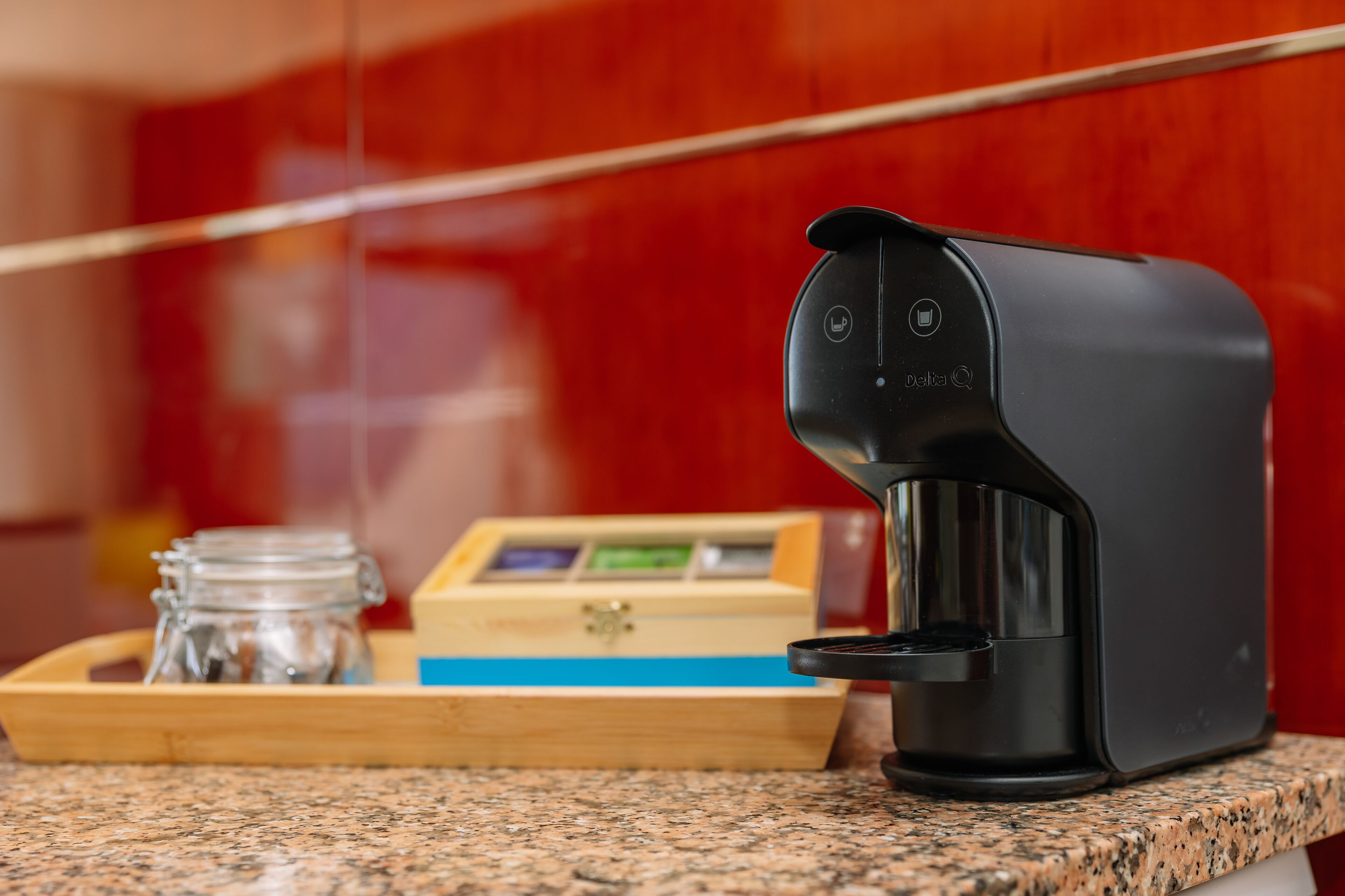 A coffee maker on a granite countertop with a tray containing a glass jar and a wooden box.
