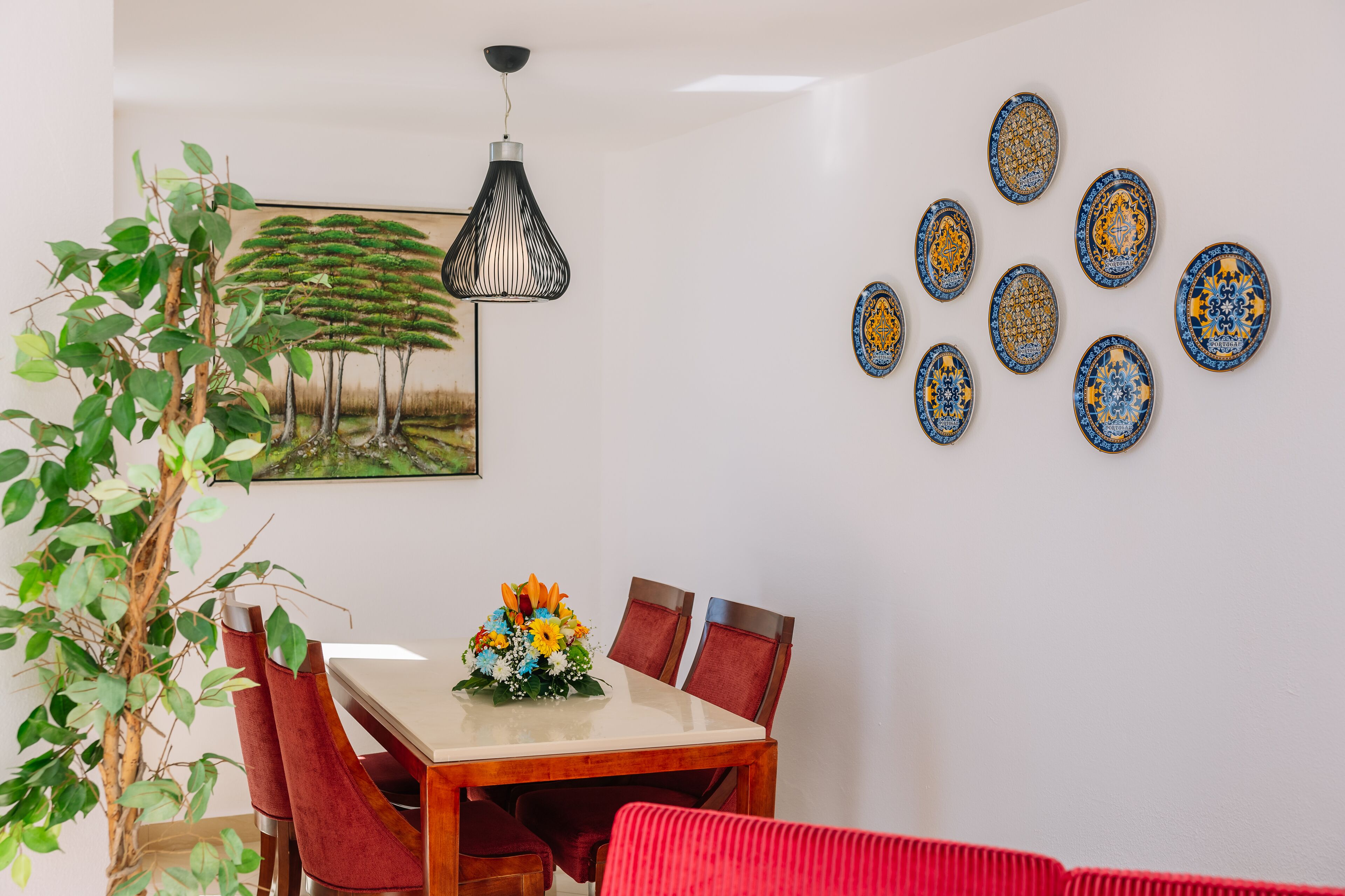 Un coin repas avec une table en bois, des chaises rouges, une plante en pot, un luminaire suspendu et un mur décoré d’assiettes.