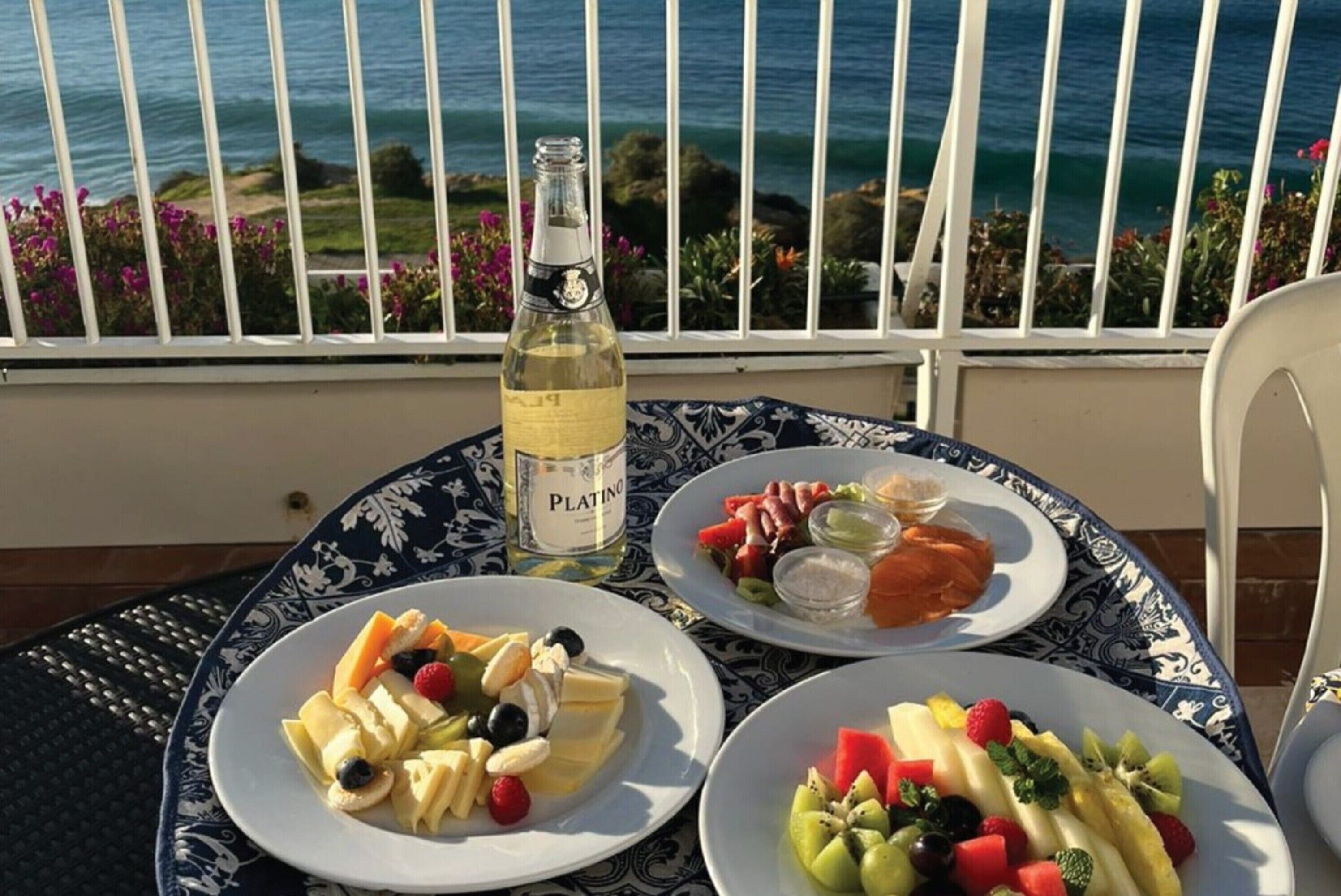 Une table garnie d’assiettes de fruits et d’une bouteille de vin Platino, le tout placé sur un balcon offrant une vue sur la mer.
