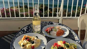 Une table garnie d’assiettes de fruits et d’une bouteille de vin Platino, le tout placé sur un balcon offrant une vue sur la mer.