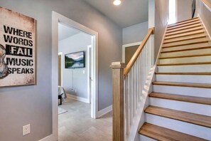 A hallway with a wooden staircase, a painting on the wall, and a television in the adjacent room.