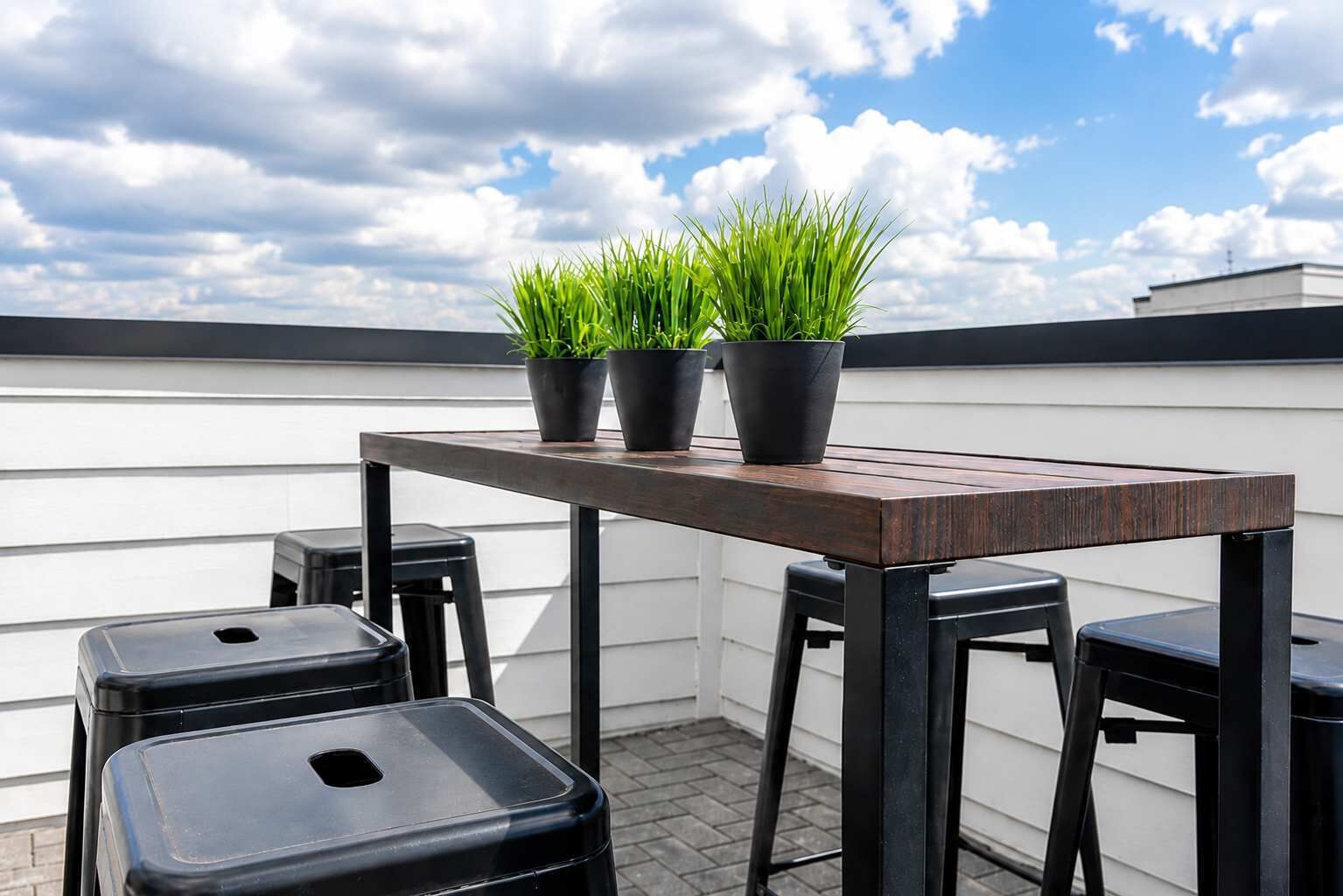 Terraço no telhado com mesa de madeira, três bancos pretos e três plantas em vasos.