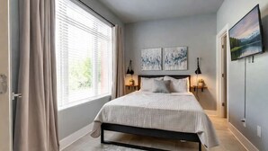 Un dormitorio con una cama, dos mesitas de noche, una ventana con cortinas y una obra de arte en la pared.