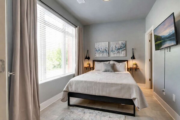 A bedroom with a bed, two bedside tables, a window with curtains, and artwork on the wall.