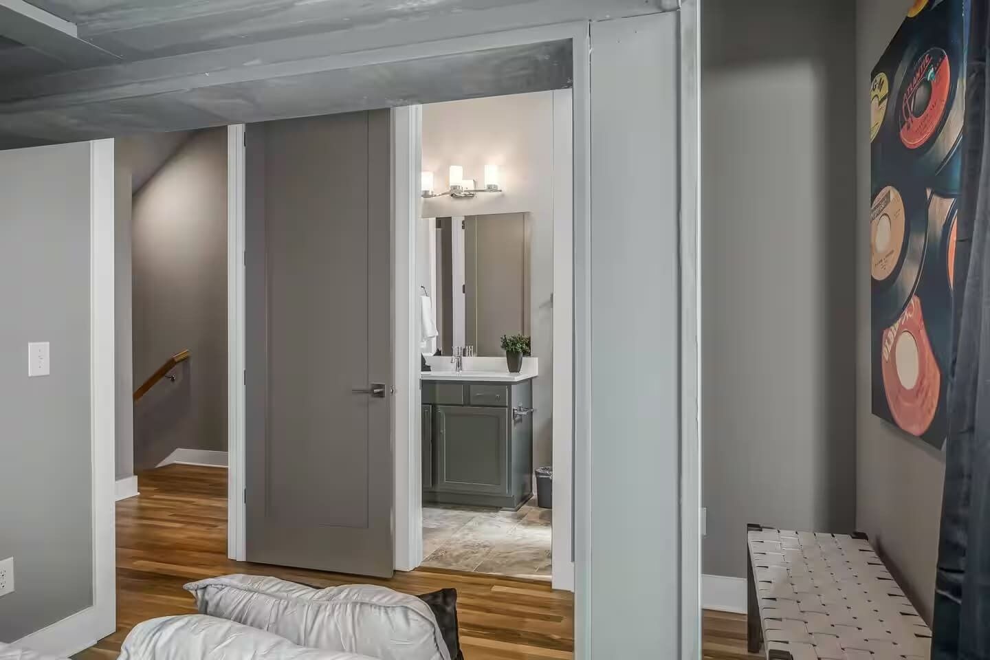 Une chambre avec un lit, une petite table de chevet et une commode. Une porte donne sur une salle de bain équipée d’un lavabo et d’un miroir. La chambre a un plancher en bois et un mur gris.