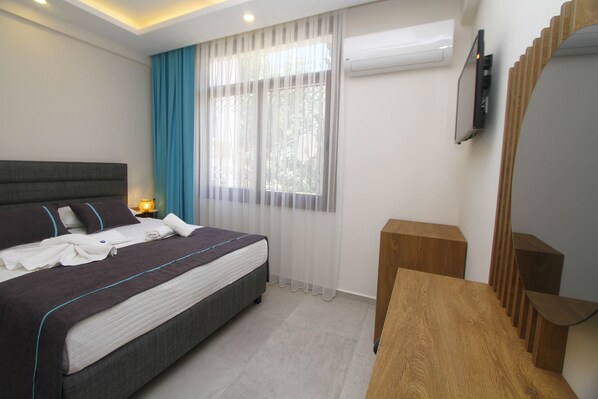 A modern bedroom with a large bed, a wooden nightstand, a TV mounted on the wall, and a window with sheer curtains.