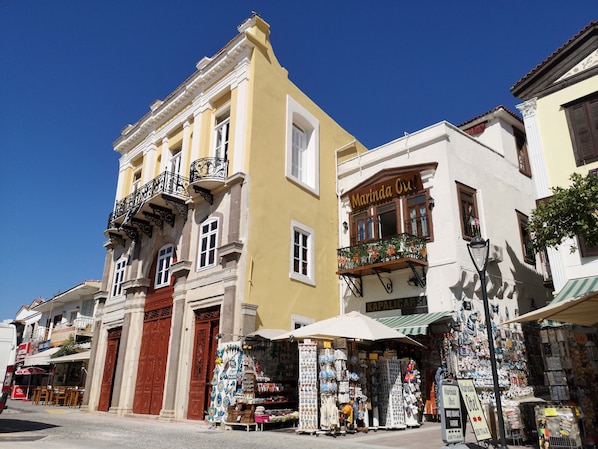 A street with a yellow building, a shop with a sign reading "Marinda Orel," and a kiosk selling various items. - Marinda Otel (Cesme)