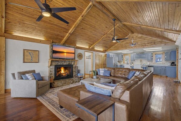 A spacious living room with a fireplace, a large sofa, a coffee table, and a kitchen in the background. - Lakefront Family Retreat • Dock • Hot Tub & Games (Cadiz)