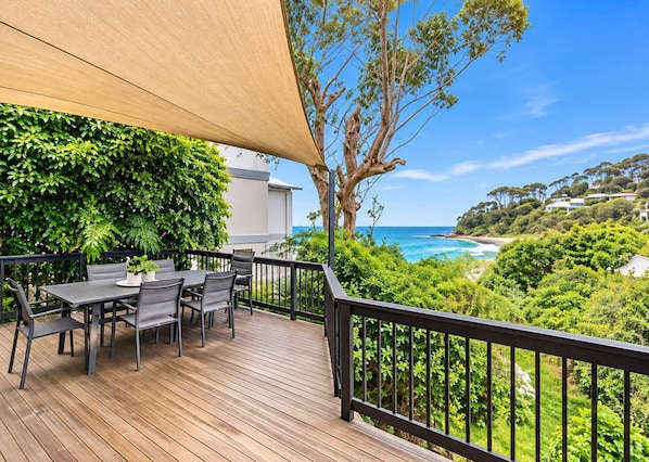 A wooden deck with a table and chairs, overlooking a coastal landscape with trees and a view of the ocean.