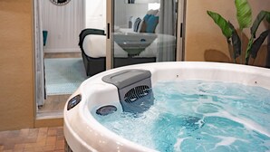 A hot tub with a control panel, a glass door leading to a room with a bed and a chair, and a plant in the background.