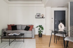 Moderni olohuone, jossa on harmaa sohva, mustia ja punaisia koristetyynyjä, lasinen sohvapöytä, ruukkukasvi sekä lasinen ruokapöytä tuoleineen.