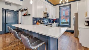 Una cocina moderna con armarios blancos, una isla grande, taburetes de bar y vistas a un edificio de ladrillo a través de una ventana.