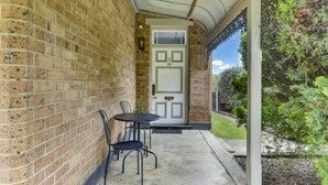A brick exterior wall with a white door, a small patio with a table and chairs, and a covered porch area.