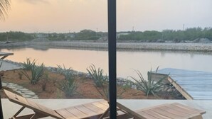 On aperçoit, à travers une porte vitrée, un espace aménagé autour d’une piscine, avec des chaises longues en bois et, au fond, une étendue d’eau calme.