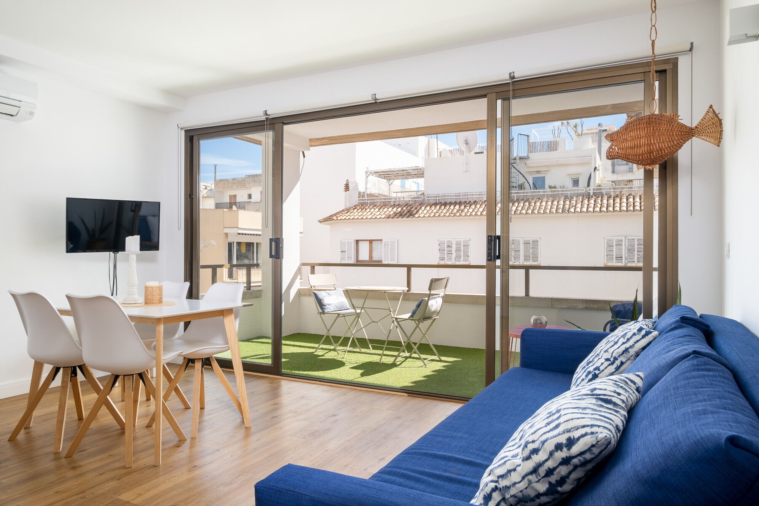 A modern living room with a large sliding glass door leading to a balcony, a blue sofa, a dining table with chairs, and a flat-screen TV.