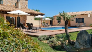 Un área junto a la piscina con sillones de descanso y sombrillas, un edificio de piedra y una palmera.