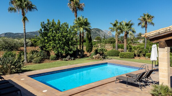 A rectangular swimming pool with a tiled edge, surrounded by a patio area with lounge chairs and a white umbrella.