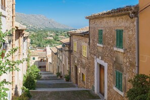 A narrow street with stone buildings, green shutters, and a mountain in the background.