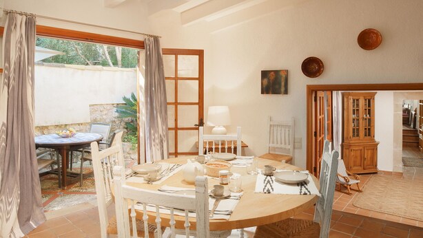 Un comedor con una mesa redonda dispuesta para cuatro personas, rodeada de sillas blancas. Se ve un aparador de madera y una pequeña mesa con una silla al fondo.