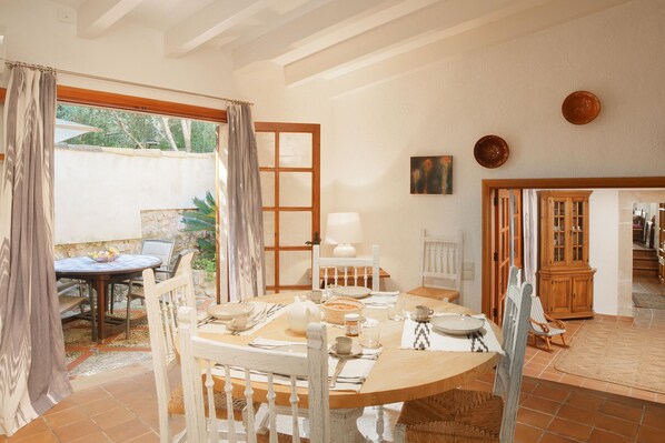 A dining area with a round table set for four, surrounded by white chairs. A wooden cabinet and a small table with a chair are visible in the background.
