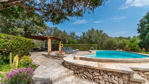 Una piscina de piedra rodeada por una terraza de piedra, una pérgola con asientos y abundante follaje.