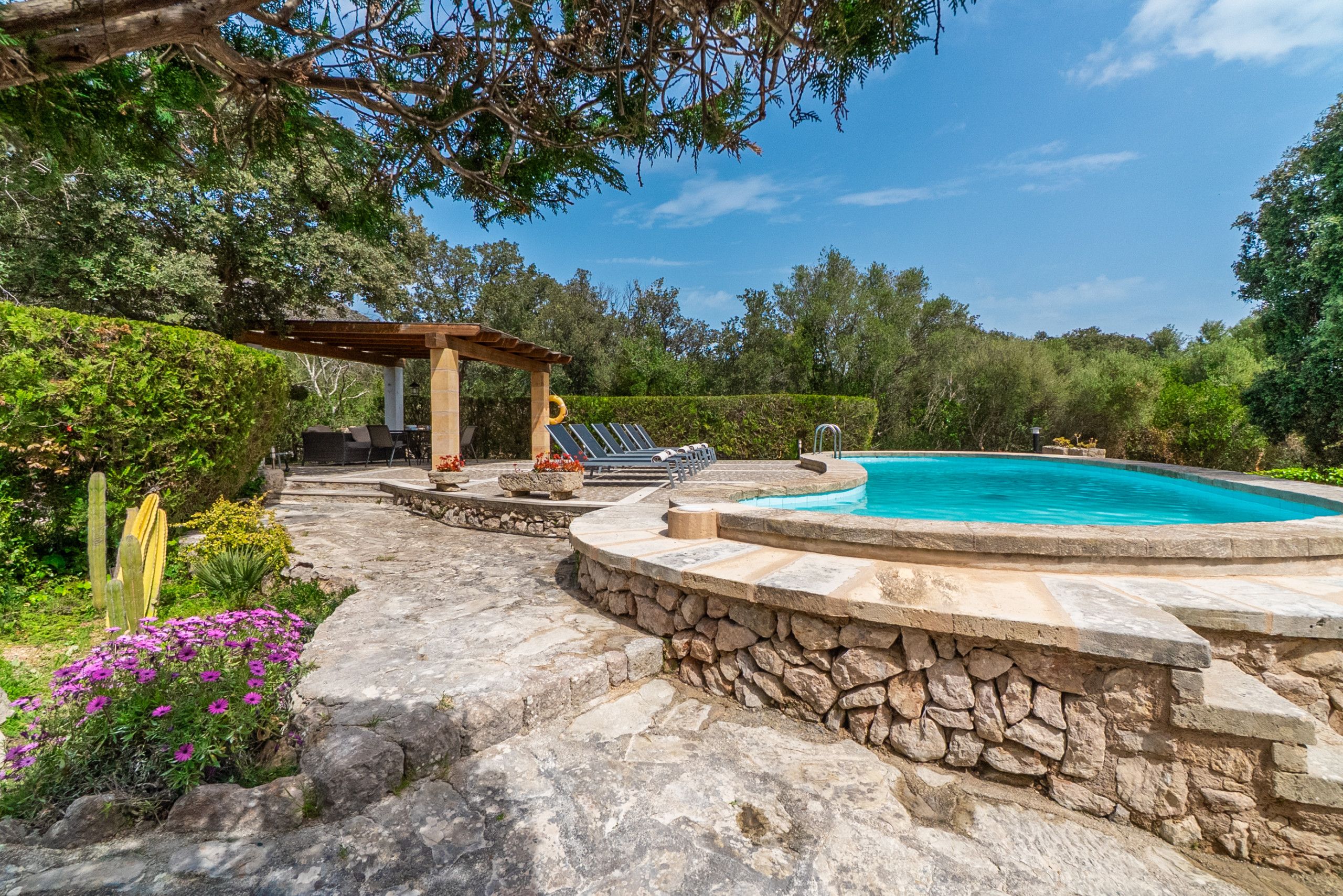 A stone pool surrounded by a stone deck, a pergola with seating, and lush greenery.