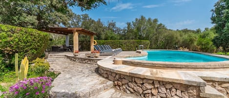 A stone pool surrounded by a stone deck, a pergola with seating, and lush greenery.