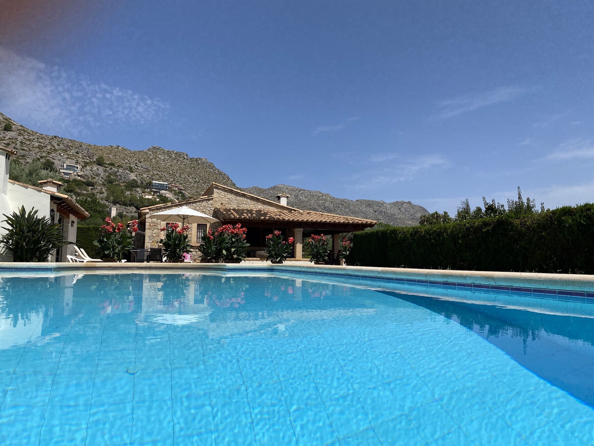 A swimming pool with clear blue water, surrounded by a building with a stone facade and a patio area with umbrellas.