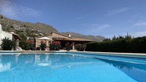 Uma piscina com água azul cristalina, rodeada por um edifício de fachada em pedra e um espaço ao ar livre com guarda-sóis.