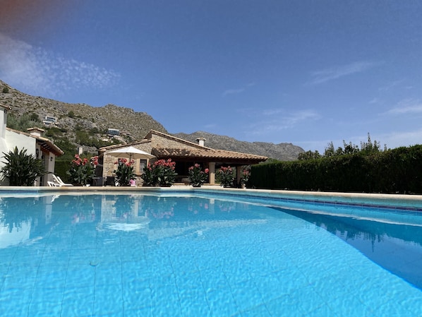 A swimming pool with clear blue water, surrounded by a building with a stone facade and a patio area with umbrellas. - Majorca holidayhome - La Font (Pollensa / Pollença)
