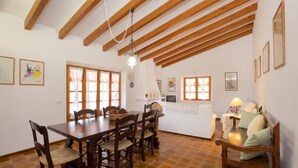 Uma área de refeições com mesa e cadeiras de madeira, uma cozinha com bancada branca e uma sala de estar com sofá e luminária.