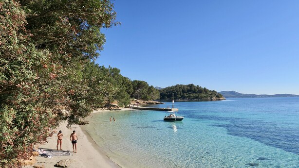 Uma praia com águas cristalinas, um barco e uma ilha arborizada ao longe.