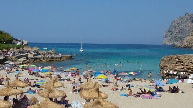Una playa concurrida con sombrillas, gente y un velero.