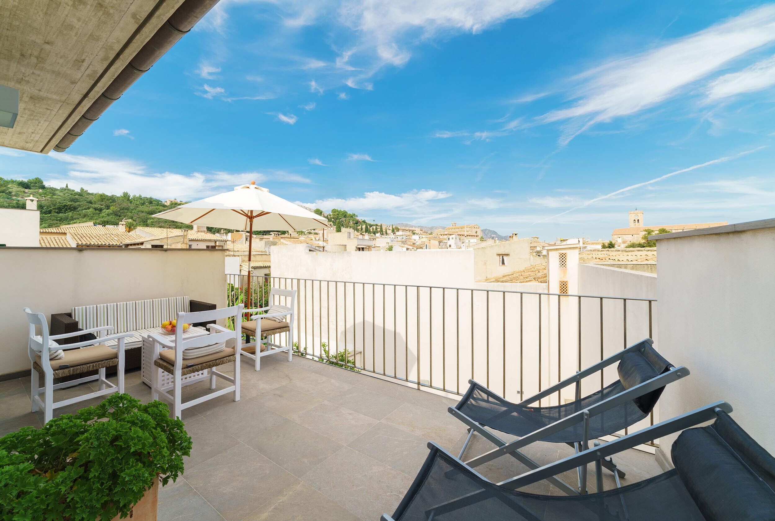 Ein Balkon mit einer Couch, Stühlen und einem Tisch, mit Blick auf eine Stadtlandschaft mit Gebäuden und einem Hügel.