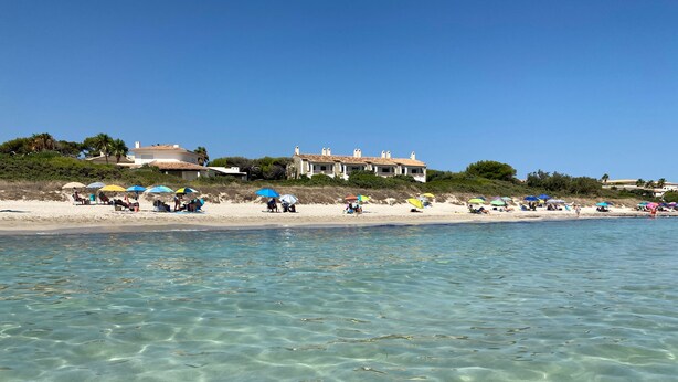 En strand med klart, grunt vatten, parasoller och människor som kopplar av.