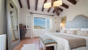A bedroom with a large bed, a bench, a window with curtains, a mirror, and a ceiling with exposed beams. - Majorca Villa with pool in Majorca - Villa Font (Mancor de la Vall)