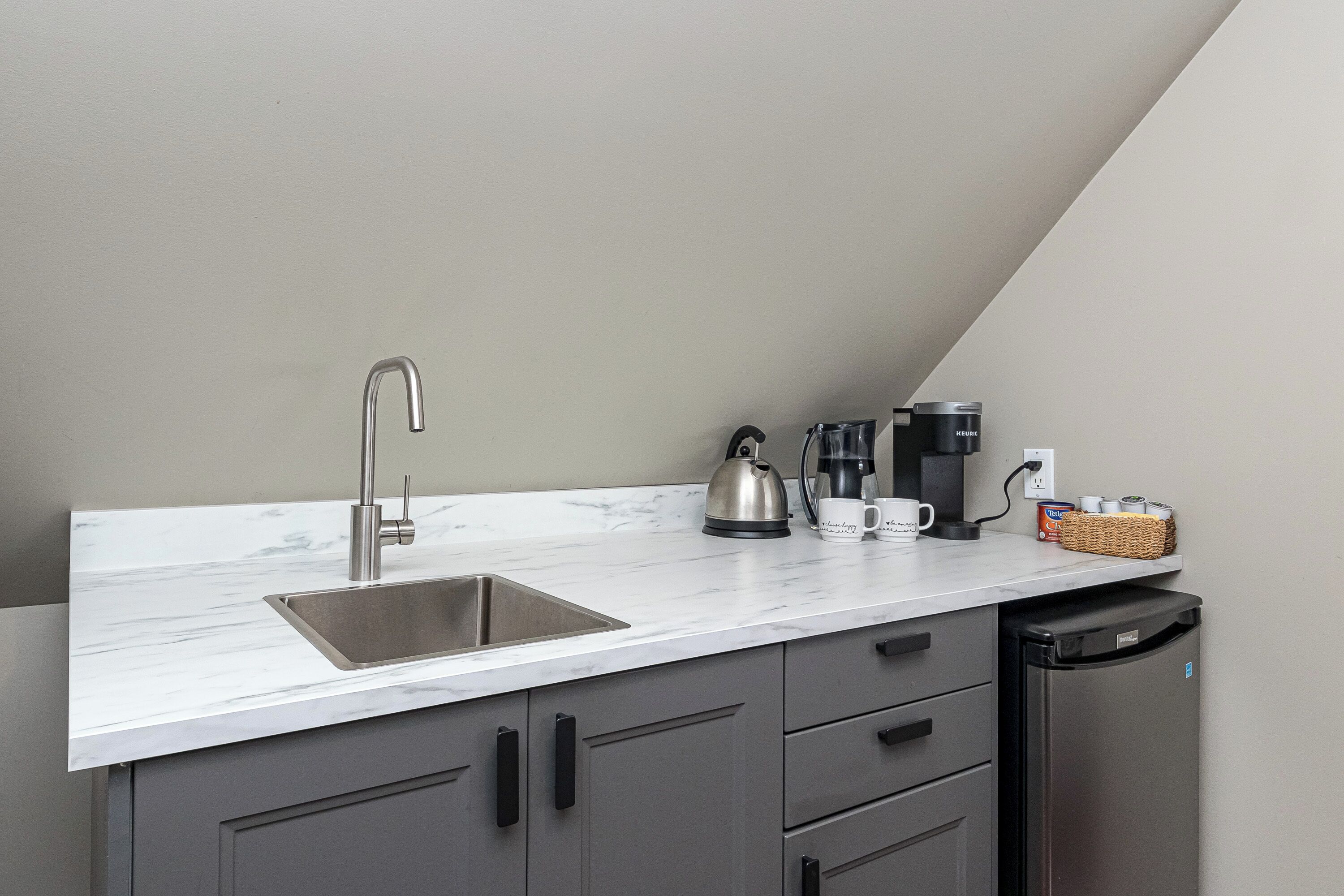 A kitchen with a sink, kettle, coffee maker, and a basket of items on the counter.